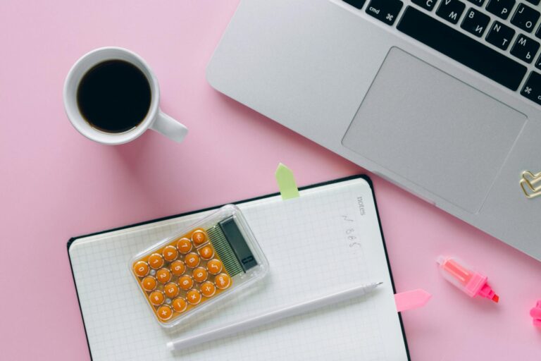 A vibrant flat lay workspace featuring a laptop, coffee, calculator, and notebook on a pink surface.