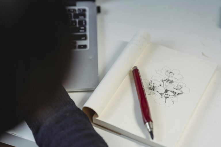 Artistic floral sketch in notebook with red pen on desk beside a laptop.